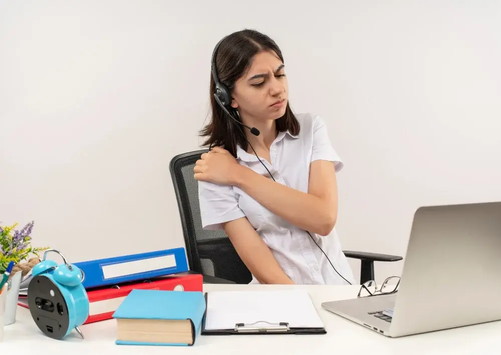young woman white shirt headphones with microphone looking unwell touching her shoulder feeling pain sitting table with folders laptop white wall
